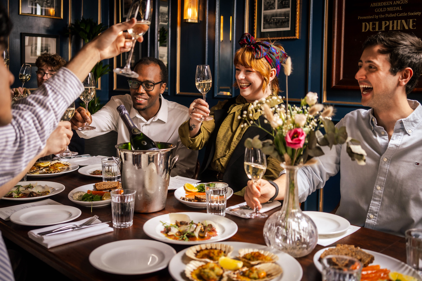 A group of friend gathered round a food laden table, raising a toast and smiling