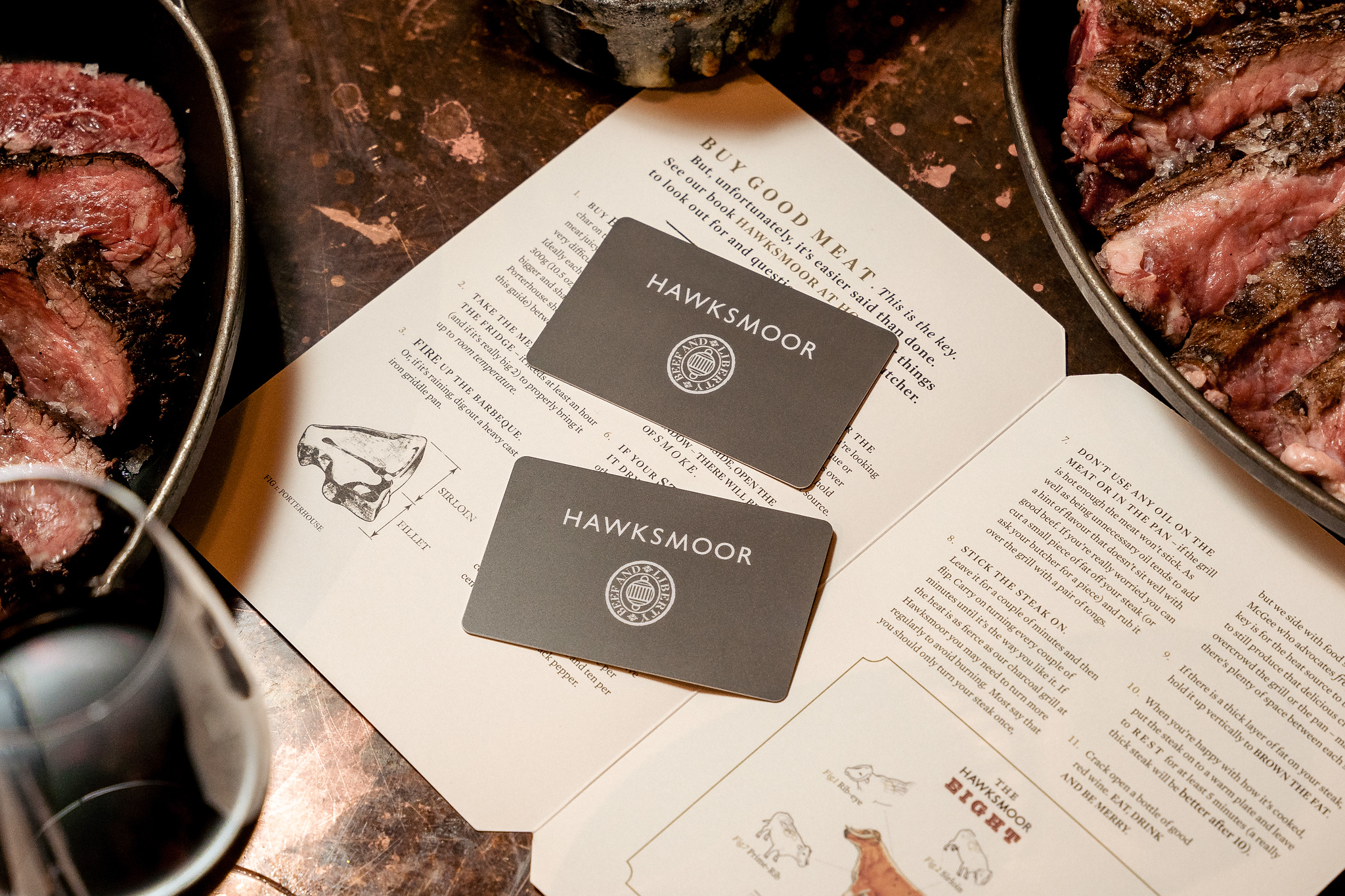 Christmas 22 Two Hawksmoor gift cards on an open Hawksmoor books, with skillets of steaks in the corner